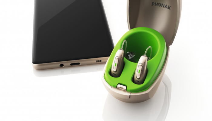 Phonak: Mini-Ladestation mit eingesteckten Hörgeräten neben Smartphone