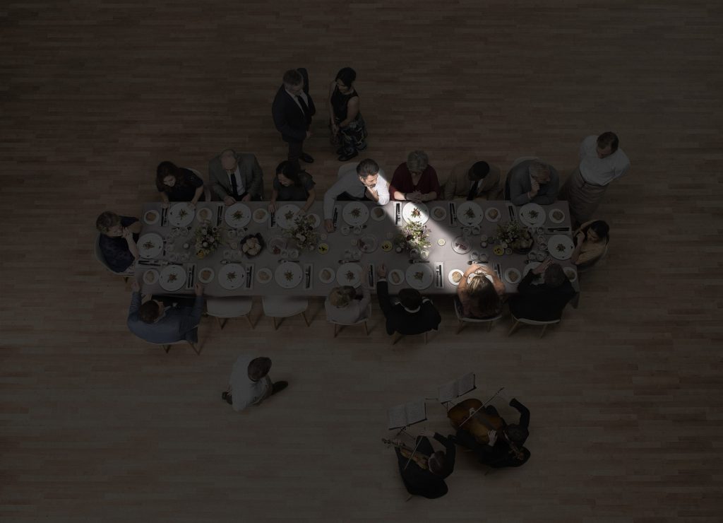 Blick von oben auf einen Tisch mit vielen Menschen. Ein Zuhörer schaut schräg gegenüber einen Gesprächspartner an. Die Ausrichtung der Richtmikrofone wird durch ein Spotlight symbolisiert.